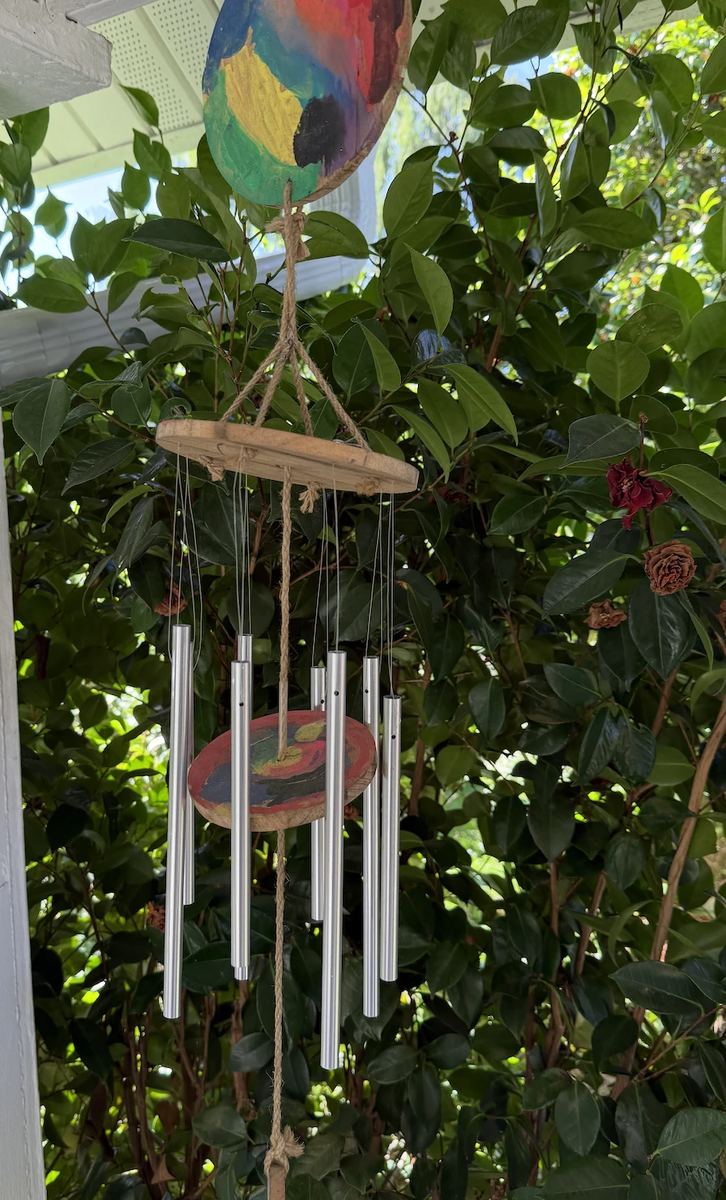 A wind chime with a painted wooden disc, hanging among leaves on a porch